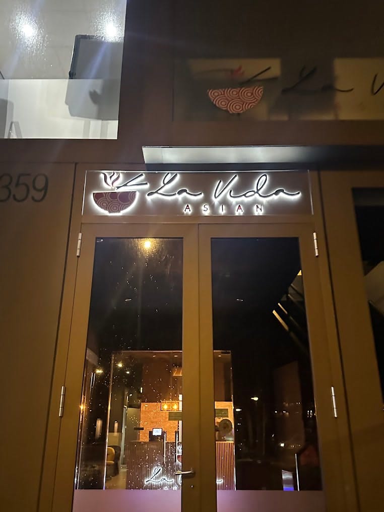 Ingang van La Vida Asian bij avond, met het verlichte logo boven de glazen deur en huisnummer 359