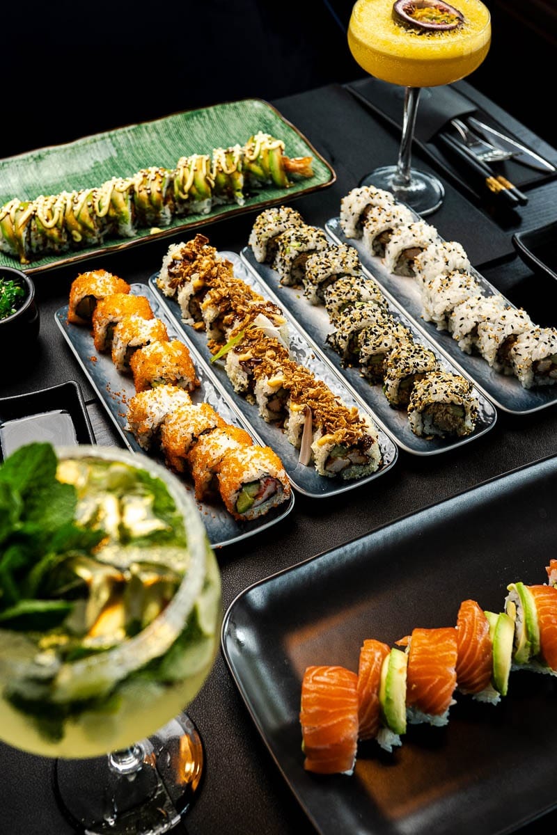 Verschillende sushirolls op lange zwarte schalen, met een passievruchtcocktail en een cocktail met munt